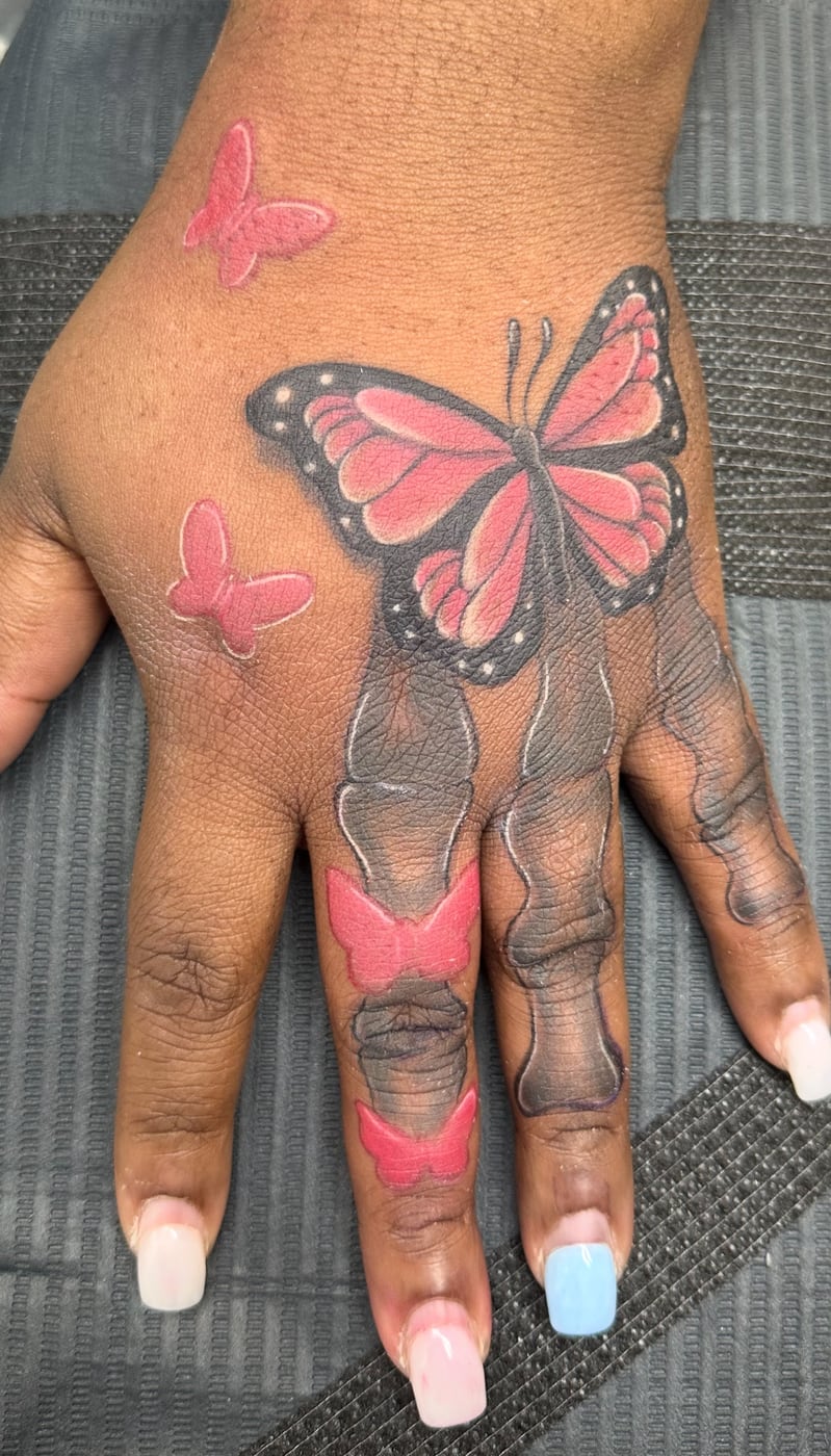 Pink butterfly tattooed across the back of a hand with smaller butterflies above it and shaded panels running down the fingers