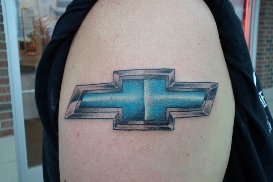 Realistic shoulder tattoo of a chrome-edged automotive bowtie badge with a brushed blue centre and cast shadow