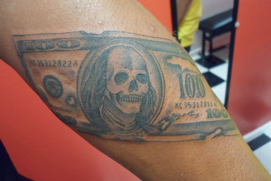 Black and grey inner arm tattoo of a hundred dollar bill with the portrait reworked as a skull, engraved detail throughout