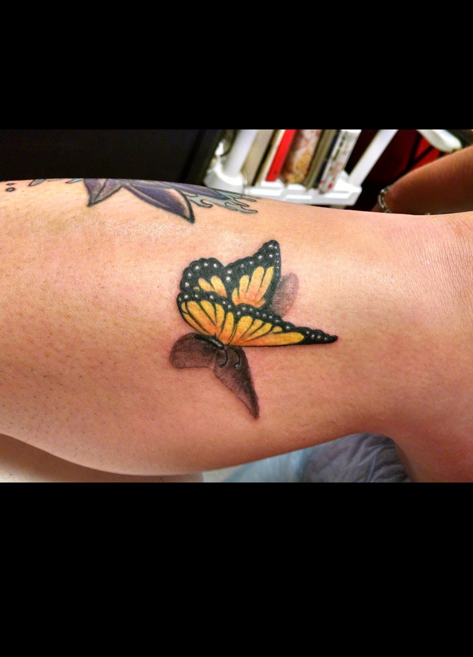Color tattoo of a monarch butterfly on an upper arm, wings rendered with a cast shadow so it lifts off the skin