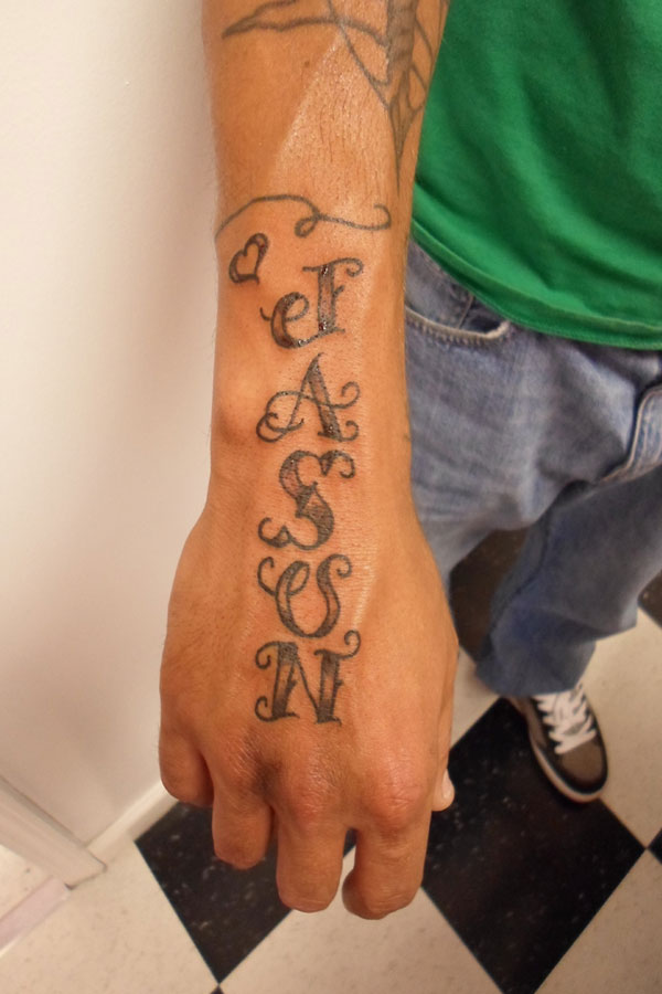 Ornate serif lettering tattooed vertically down the side of the hand and wrist, each letter drawn with decorative flourishes