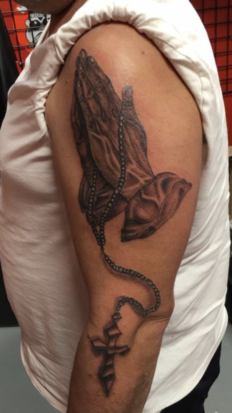 Black and grey tattoo of praying hands holding a rosary, the beads trailing down the arm to a cross below the elbow