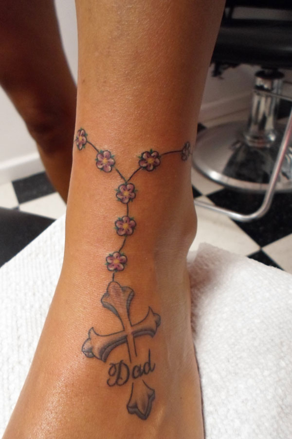 Memorial ankle tattoo of a rosary made from small pink flowers leading down to a cross lettered with the word Dad