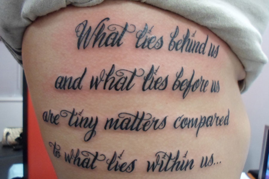 Four lines of ornate black calligraphy script tattooed across the ribcage, hand-drawn to follow the curve of the body