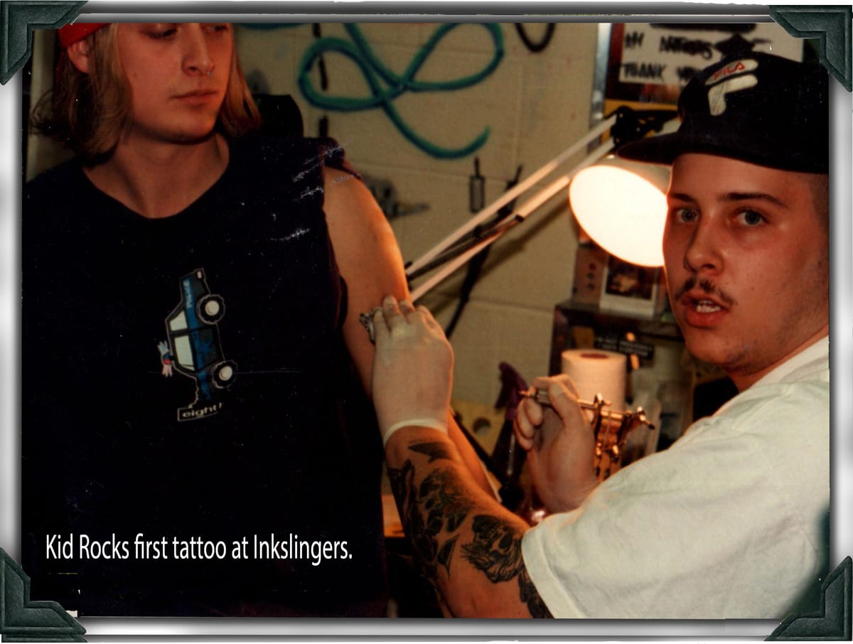 A tattooer working on a seated client's arm in a busy studio in the 1990s, both looking toward the camera