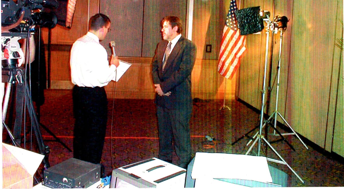 Mike Jeziak Sr. being interviewed under television lights in a wood-panelled room in Washington D.C., an American flag behind him