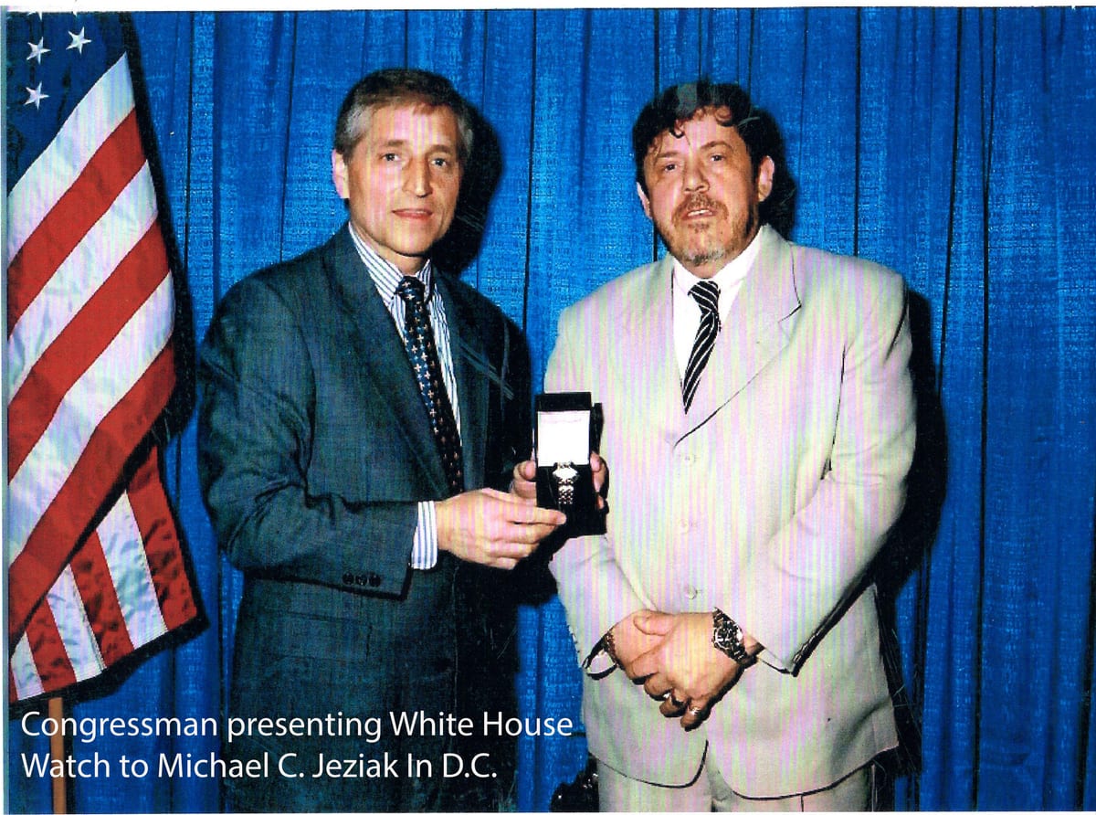A congressman presenting a cased White House watch to Michael C. Jeziak in front of an American flag and blue curtain